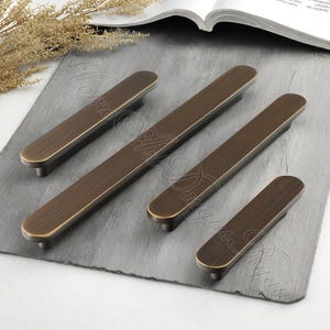 K&ouml;nnte beinhalten: Ein Satz von vier bronzefarbenen Schrankgriffen mit rechteckigem Design. Die Griffe haben eine geb&uuml;rstete Oberfl&auml;che und abgerundete Enden. Die Hardware wird auf einer grauen Oberfl&auml;che pr&auml;sentiert, die die verschiedenen Gr&ouml;&szlig;en zeigt.