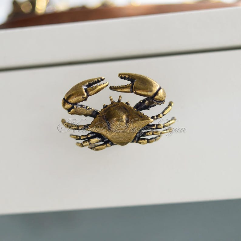 Puede incluir: Un tirador de caj&oacute;n en forma de cangrejo de color dorado. El cangrejo est&aacute; detallado con patas y garras texturizadas.