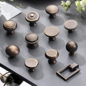 K&ouml;nnte beinhalten: Eine Auswahl an bronzefarbenen Schrankkn&ouml;pfen und -griffen in verschiedenen Formen. Die Hardware umfasst runde, flache und kugelf&ouml;rmige Kn&ouml;pfe sowie einen rechteckigen Griff. Die Oberfl&auml;che hat ein Vintage- und Antik-Aussehen.