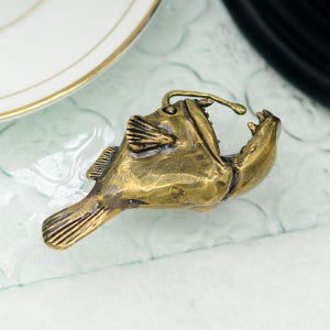 K&ouml;nnte beinhalten: Bronze-farbene Anglerfisch-Figur mit offenem Maul, sichtbaren Z&auml;hnen und detaillierten Flossen. Die Figur hat eine Schlaufe oben, m&ouml;glicherweise als Anh&auml;nger. Das Design ist detailliert und strukturiert.