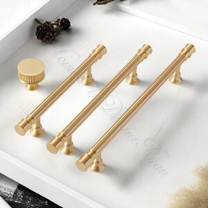 Puede incluir: Un conjunto de herrajes dorados para armarios, que incluye tres tiradores largos y un pomo redondo. Los tiradores tienen un dise&ntilde;o acanalado y se muestran sobre una superficie blanca. Adecuado para armarios de cocina o ba&ntilde;o.