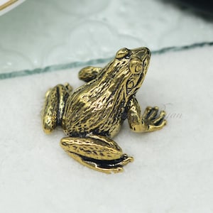 May include: A small, gold-colored frog figurine. The frog is sitting on a white surface.