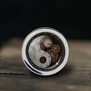 May include: A silver-colored round pendant with a yin yang symbol design featuring intricate gears and cogs in a copper color.
