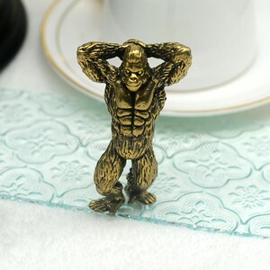 Op de afbeelding: Een bronzen gorilla beeldje met de handen achter het hoofd. De gorilla heeft een gespierd lichaam en staat rechtop. Het beeldje is gedetailleerd met getextureerde vacht en gelaatstrekken. De achtergrond is wazig, met een vleugje van een witte kop.