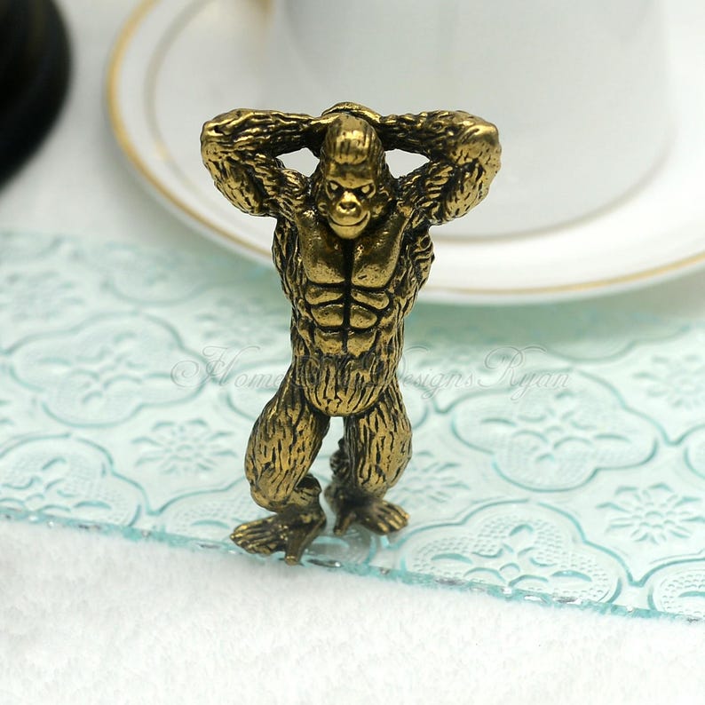 Op de afbeelding: Een bronzen gorilla beeldje met de handen achter het hoofd. De gorilla heeft een gespierd lichaam en staat rechtop. Het beeldje is gedetailleerd met getextureerde vacht en gelaatstrekken. De achtergrond is wazig, met een vleugje van een witte kop.