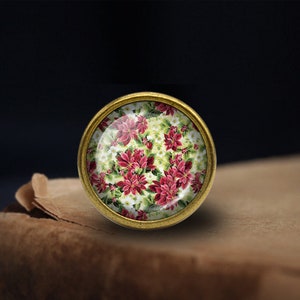 May include: A round, gold-toned, decorative knob with a glass top featuring a red and white floral pattern. The floral pattern is reminiscent of poinsettias.
