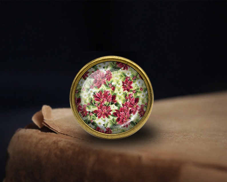 May include: A round, gold-toned, decorative knob with a glass top featuring a red and white floral pattern. The floral pattern is reminiscent of poinsettias.