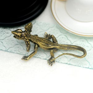 Peut inclure: Une figurine en métal de couleur dorée représentant un lézard avec une longue queue et de grandes oreilles pointues. Le lézard est en position de marche, la tête tournée sur le côté.