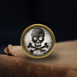 May include: A round, gold-toned metal pin with a glass cabochon featuring a black skull and crossbones design on a white background.