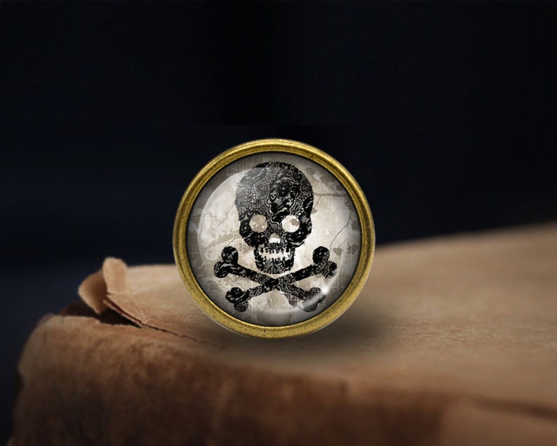 May include: A round, gold-toned metal pin with a glass cabochon featuring a black skull and crossbones design on a white background.