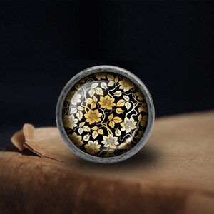 May include: A silver knob with a round glass top featuring a black and gold floral pattern.