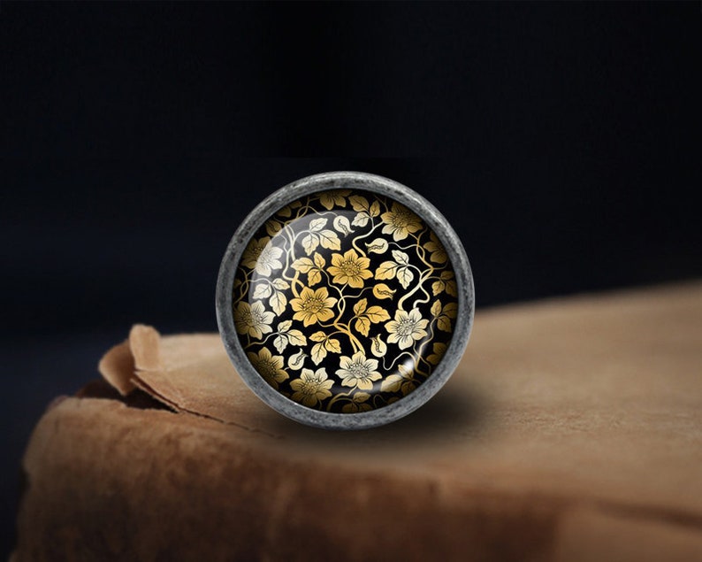 May include: A silver knob with a round glass top featuring a black and gold floral pattern.