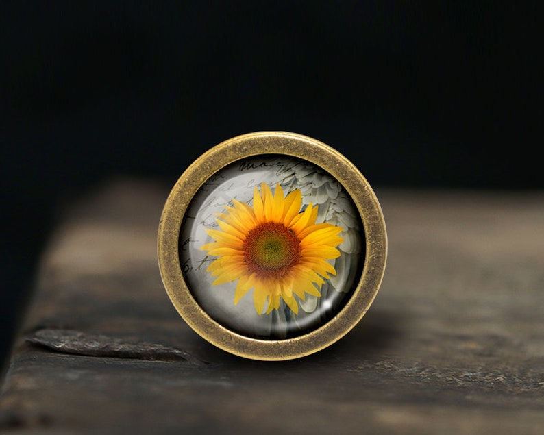 May include: A round, brass-colored pendant with a glass dome featuring a yellow sunflower with a brown center. The sunflower is on a white background with faded writing.
