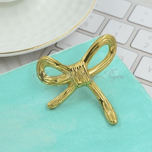 May include: Gold-toned metal bow tie shaped drawer pull. The bow is tied in a knot and has a textured surface.