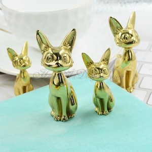 Pode incluir: Três estatuetas de gato douradas de tamanhos diferentes. O gato maior está sentado com as pernas enfiadas por baixo. Os dois gatos menores estão em pé. Os gatos têm olhos grandes e redondos e uma aparência estilizada e caricata.