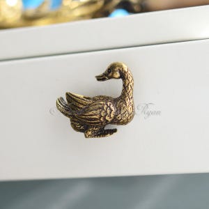 Op de afbeelding: Een gouden eenden-ladegreep met een gedetailleerd ontwerp. De eend kijkt naar rechts en heeft zijn vleugels uitgespreid. De greep is gemonteerd op een wit oppervlak.