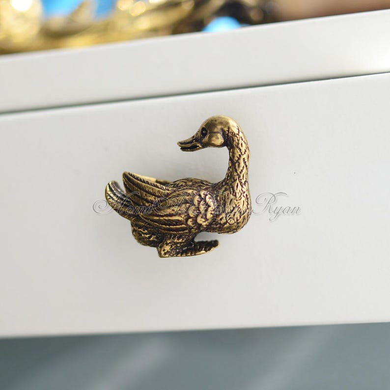 Op de afbeelding: Een gouden eenden-ladegreep met een gedetailleerd ontwerp. De eend kijkt naar rechts en heeft zijn vleugels uitgespreid. De greep is gemonteerd op een wit oppervlak.