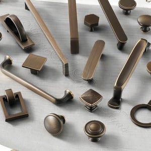 K&ouml;nnte beinhalten: Eine Auswahl an bronzefarbener Schrankbeschl&auml;ge, darunter Kn&ouml;pfe, Griffe und Ziehgriffe. Die Beschl&auml;ge weisen verschiedene Formen auf, wie z. B. runde, quadratische und rechteckige Designs. Die Beschl&auml;ge werden auf einer hellgrauen Oberfl&auml;che pr&auml;sentiert.