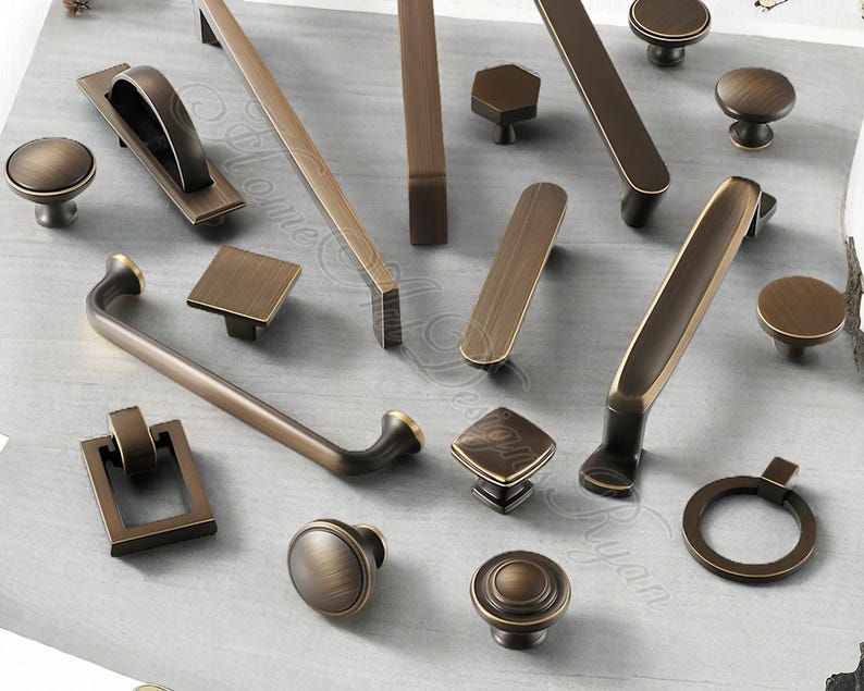 K&ouml;nnte beinhalten: Eine Auswahl an bronzefarbener Schrankbeschl&auml;ge, darunter Kn&ouml;pfe, Griffe und Ziehgriffe. Die Beschl&auml;ge weisen verschiedene Formen auf, wie z. B. runde, quadratische und rechteckige Designs. Die Beschl&auml;ge werden auf einer hellgrauen Oberfl&auml;che pr&auml;sentiert.