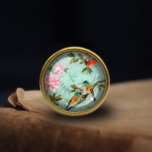 May include: A round, gold-toned metal knob with a glass insert featuring a painted design of birds and flowers on a light blue background.