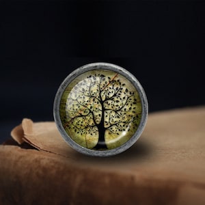 May include: A silver knob with a glass insert featuring a black silhouette of a tree with green leaves on a yellow background.