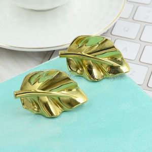 Könnte beinhalten: Ein Paar goldene Ohrringe in Blattform. Die Ohrringe sind aus glänzendem Metall gefertigt und haben ein detailliertes Blattdesign.