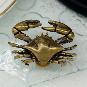 Puede incluir: Un pomo de armario o tirador de caj&oacute;n de lat&oacute;n con forma de cangrejo. El cangrejo est&aacute; detallado con garras, patas y una concha texturizada.