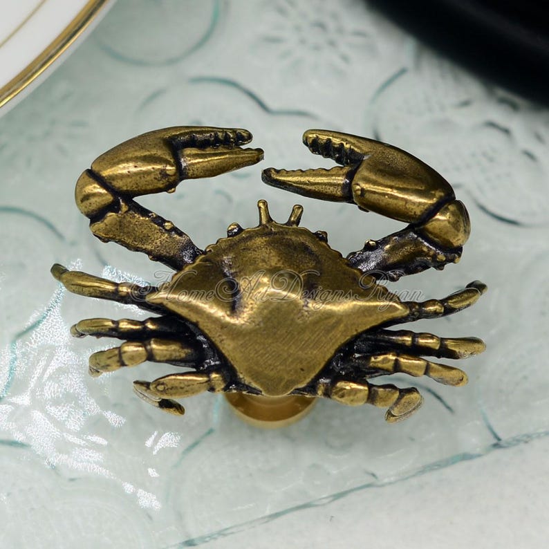 Puede incluir: Un pomo de armario o tirador de caj&oacute;n de lat&oacute;n con forma de cangrejo. El cangrejo est&aacute; detallado con garras, patas y una concha texturizada.