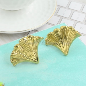 May include: A pair of gold-tone earrings shaped like a fan. The earrings are made of metal and have a textured surface.