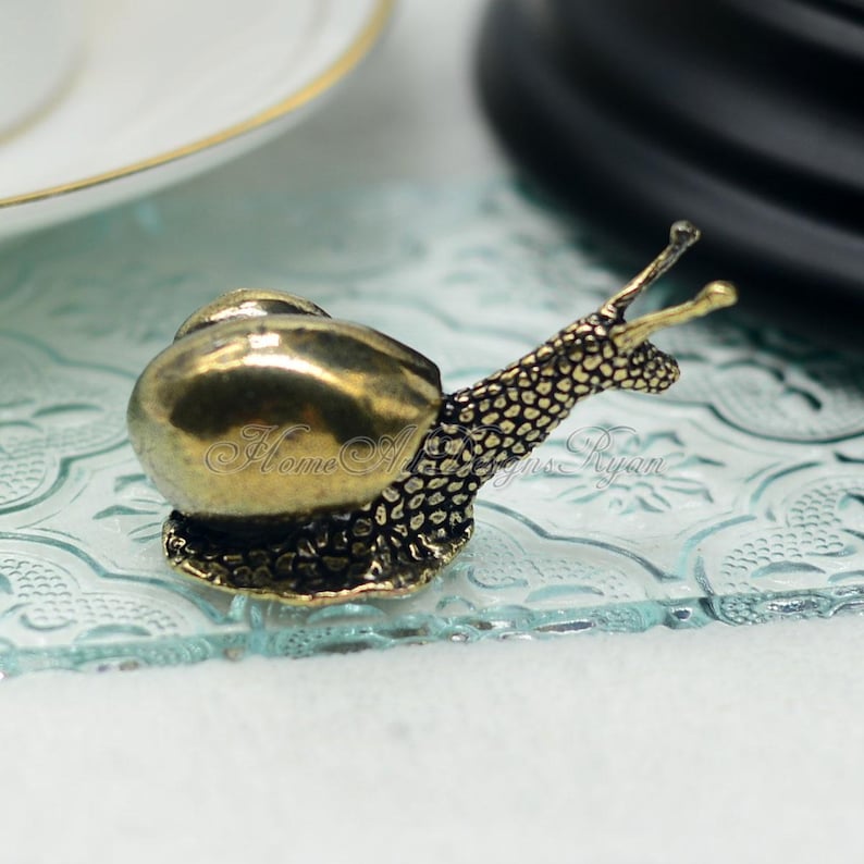 Puede incluir: Una peque&ntilde;a figurita de caracol de color dorado con una concha detallada y un cuerpo texturizado. El caracol est&aacute; sentado sobre una superficie de vidrio transparente con un patr&oacute;n decorativo.