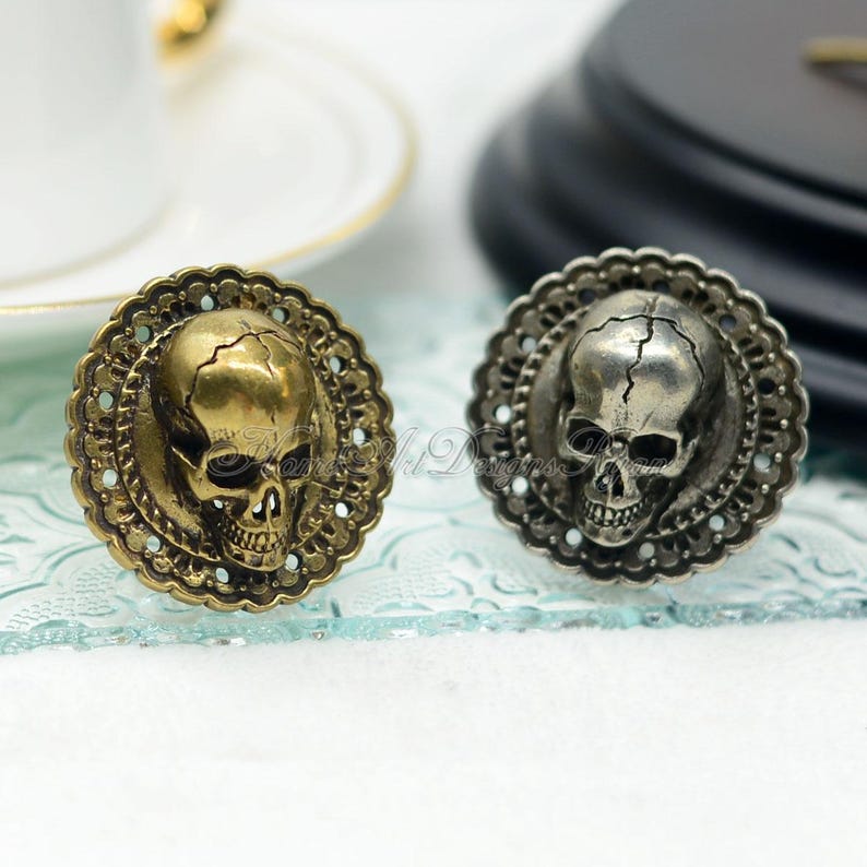 May include: Two decorative skull-shaped cabinet knobs. One is gold-toned, the other is silver-toned. Each skull is centered within a scalloped, ornate frame. The knobs are likely made of metal and are designed for furniture or decorative use.