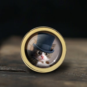 Puede incluir: Un marco redondo dorado con una lente de vidrio que muestra un gato con un sombrero de copa negro y gafas redondas. El gato está mirando directamente al espectador.