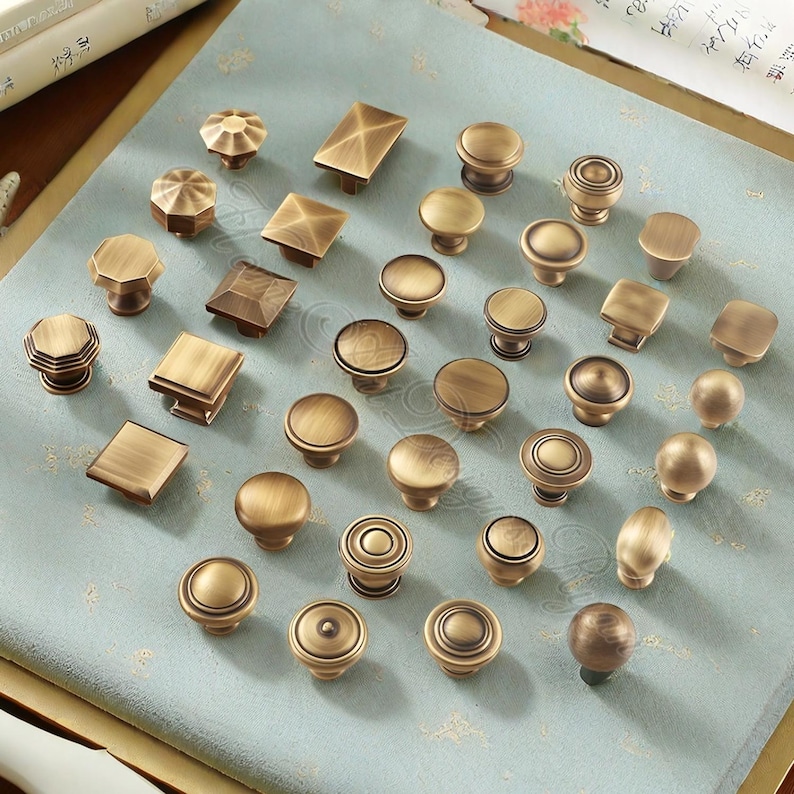 May include: A selection of antique brass cabinet knobs in diverse shapes and designs. The knobs feature round, square, and faceted styles, all with a warm, aged finish. They are arranged on a light blue patterned surface.