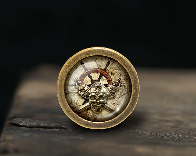 May include: A round, brass-coloured pendant with a glass insert featuring a pirate skull wearing a hat, crossed swords, a compass, and a ship's wheel on a vintage map background.