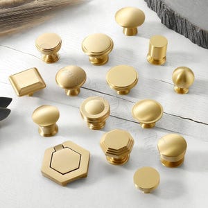 Puede incluir: Una colecci&oacute;n de diversos pomos de lat&oacute;n para armarios en diferentes formas, incluyendo dise&ntilde;os redondos, cuadrados y hexagonales. Los pomos tienen un acabado cepillado y est&aacute;n dispuestos sobre una superficie clara.