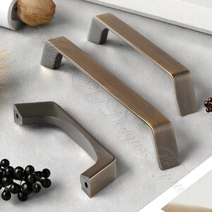 K&ouml;nnte beinhalten: Drei Metall-Schrankgriffe in Bronze- und Graut&ouml;nen. Die Griffe haben eine rechteckige Form mit einem gebogenen Griff. Die Hardware ist f&uuml;r Schubladen oder Schr&auml;nke konzipiert. Die Griffe werden auf einer hellgrauen Oberfl&auml;che gezeigt.