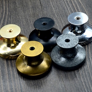 May include: Five different cabinet knobs in various finishes. The knobs are round with a raised centre and a smooth surface. The finishes include gold, brass, black, silver, and grey.