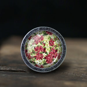 May include: A round, silver-colored cabinet knob with a floral design. The design features red poinsettia flowers and green leaves on a white background.