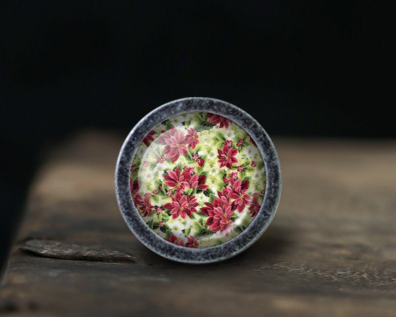 May include: A round, silver-coloured cabinet knob with a floral design. The design features red poinsettia flowers and green leaves on a white background.