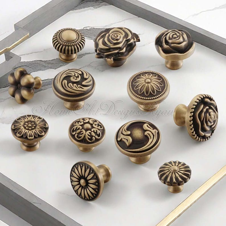 Pu&ograve; includere: Un assortimento di pomelli per mobili in ottone antico con vari disegni floreali e ornati. I pomelli presentano motivi dettagliati, tra cui rose, girasoli e motivi a spirale. I pomelli sono esposti su una superficie di marmo bianco.