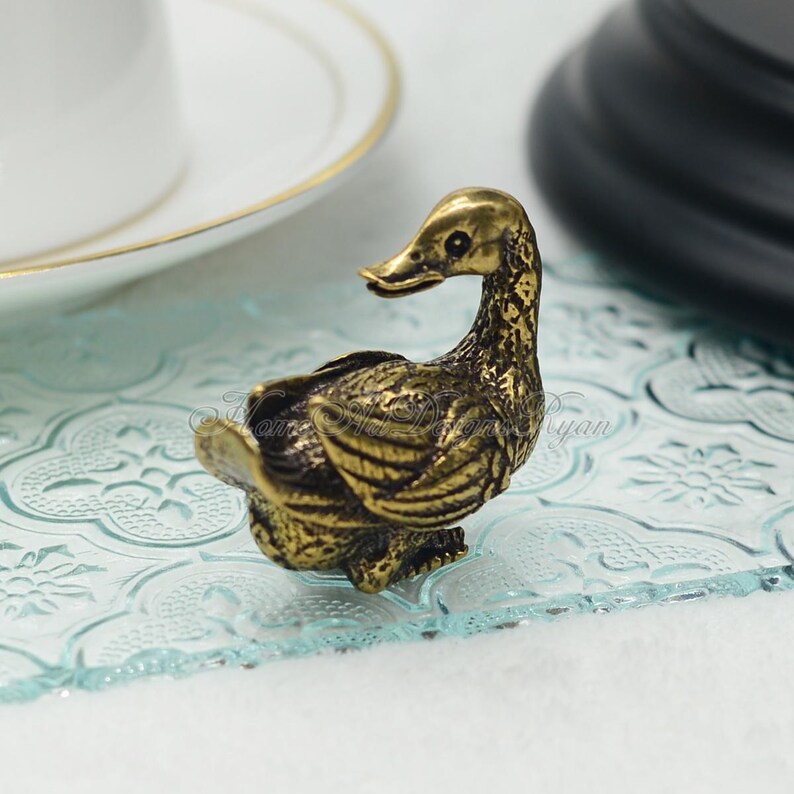 Op de afbeelding: Een klein, goudkleurig eendje met ingewikkelde details. Het eendje zit op een lichtblauw, gepatroneerd oppervlak.