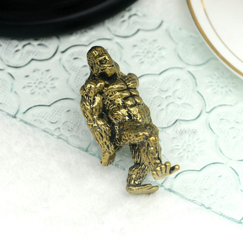 K&ouml;nnte beinhalten: Eine goldfarbene Metallfigur eines Gorillas, der mit gekreuzten Armen sitzt. Die Figur ist detailliert mit Muskeldefinition und hat eine strukturierte Oberfl&auml;che.