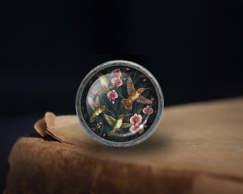 May include: A silver knob with a round glass insert featuring a floral pattern with hummingbirds in shades of green, pink, and orange.