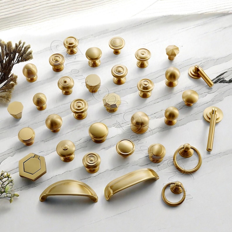 Puede incluir: Una colecci&oacute;n de pomos y tiradores de lat&oacute;n para armarios de diferentes formas y tama&ntilde;os. Los herrajes incluyen dise&ntilde;os redondos, hexagonales y en forma de barra, todos con un acabado dorado mate. Los pomos y tiradores est&aacute;n dispuestos sobre una superficie blanca.