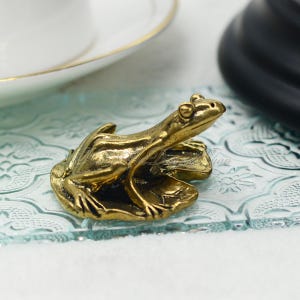 以下が含まれることがあります： 葉の上に座っている金色のカエルの置物。細部まで丁寧に作られており、リアルな見た目です。