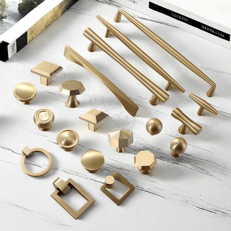 Puede incluir: Una colecci&oacute;n de herrajes dorados para gabinetes, que incluyen perillas y tiradores, en varias formas y tama&ntilde;os. Los herrajes incluyen dise&ntilde;os redondos, cuadrados y geom&eacute;tricos. Los tiradores son largos y delgados, mientras que las perillas son m&aacute;s compactas.