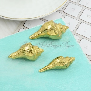 Könnte beinhalten: Drei goldfarbene Muscheln auf einer türkisfarbenen Oberfläche. Die Muscheln haben alle die Form eines Kegels, mit einer spitzen Spitze und einer breiten Öffnung an der Basis.