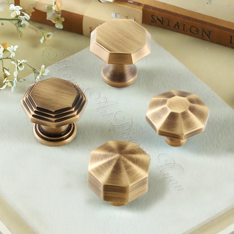 May include: Four antique brass cabinet knobs with an octagonal design. The knobs have a vintage aesthetic and are arranged on a light-coloured surface. The knobs are likely for furniture or decorative purposes.