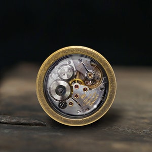 Puede incluir: Primer plano de un movimiento de reloj de color dorado con engranajes visibles y el texto "17 JEWELS SWISS  LONGINES"