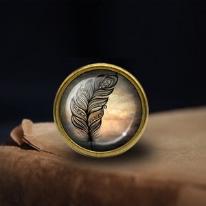 May include: A gold-toned metal pin with a glass cabochon featuring a black and white stylized feather design on a cloudy background.
